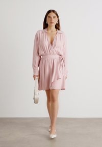 Light pink wrap dress with long sleeves, adjustable waist tie, and an asymmetric hem. Model wears white heels and holds a white handbag.