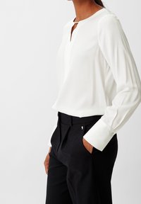 Blouse blanche avec col en V et petit détail en métal argenté, associée à un pantalon noir taille haute à design plissé.