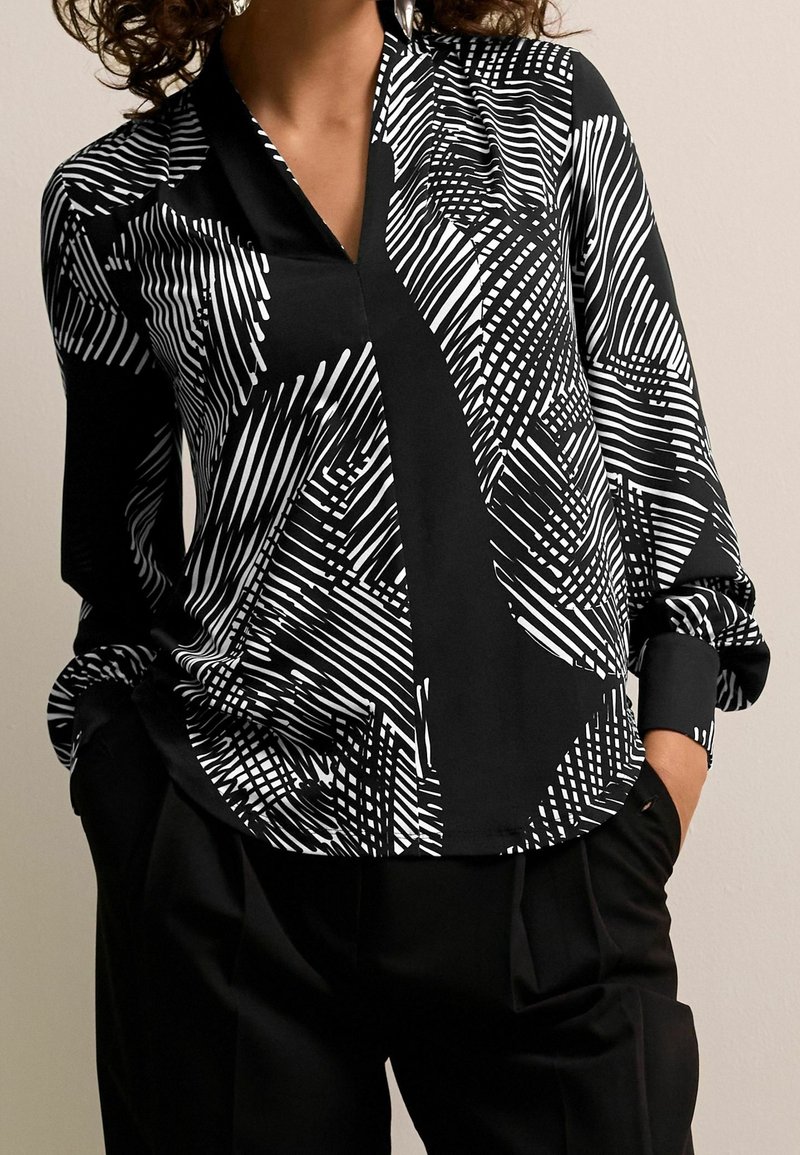Femme portant un chemisier à manches longues à motif géométrique noir et blanc avec un pantalon noir, mains dans les poches devant un fond uni.