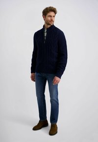 Marineblauer Kabelstrickpullover mit hohem Kragen und Reißverschluss, kombiniert mit blauen Jeans und braunen Stiefeletten. Der Pullover hat eine strukturierte Stickerei.