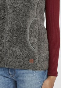 Grau gefütterte Weste mit einem Reißverschluss auf der Vorderseite, seitlichen Taschen und einem braunen Lederlogo-Patch. Über einem kastanienbraunen, gerippten Langarmshirt getragen.