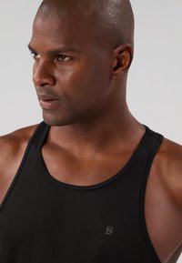 Top cropped preto com uma textura canelada, apresentando um decote redondo e um pequeno logótipo em destaque. Close-up da peça no ombro de um modelo.