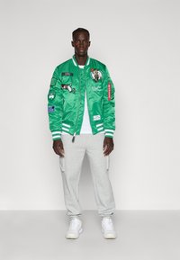 Jachetă bomber din satin verde cu logo-ul Celtics și patch-uri, accente albe, asortată cu pantaloni cargo gri și adidași albi.