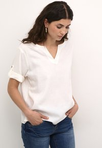 Bela lanena bluzica z V-izrezom in zavihanimi rokavi, s subtilno teksturo in ohlapnim krojem. V kombinaciji z modrimi jeans hlačami.