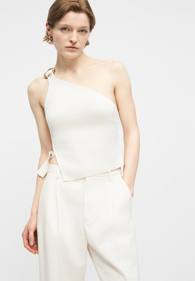 Γυναίκα με κοντά μαλλιά που φοράει λευκό τοπ ενός ώμου με χρυσές αγκράφες και λευκό φαρδύ παντελόνι, στέκεται με το ένα χέρι στην τσέπη.