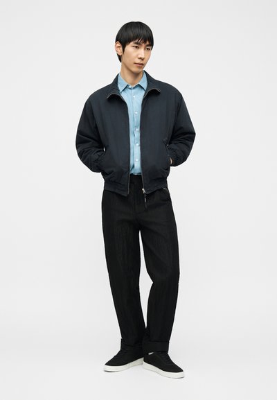 Νεαρός άνδρας που φοράει μαύρο μπουφάν πάνω από ανοιχτό μπλε πουκάμισο, μαύρο παντελόνι και μαύρα αθλητικά παπούτσια με λευκή σόλα, στέκεται με τα χέρια στις τσέπες.