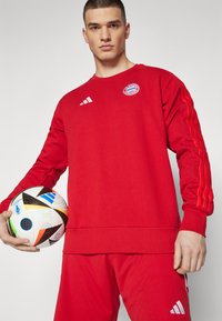 Rød genser og bukser med tre hvite striper på armene, med Bayern Munich-logo. Modellen holder en mønstrete fotball.