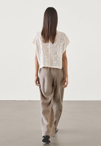 Nainen kävelee poispäin valkoisessa pitsisessä lyhythihaisessa paidassa ja löysissä ruskeissa pellavahousuissa mustilla sandaaleilla, yksinkertaista valkoista taustaa vasten.