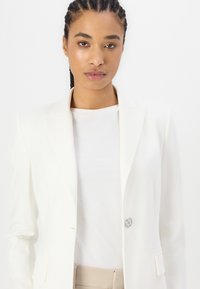 Hvit blazer med en enkelt knappåpning, noterte reverser og to frontlommer, båret over en enkel hvit skjorte og beige bukser.