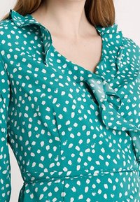 Tyrkysové šaty s bílými puntíky, s volánovým výstřihem a límečkem. Vyrobeny z měkké látky s hladkou strukturou.