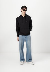 Zwarte katoenen hoodie met een voorzak, gecombineerd met lichtblauwe wijde jeans en witte sneakers. Het model draagt een paar ronde zwarte zonnebrillen.