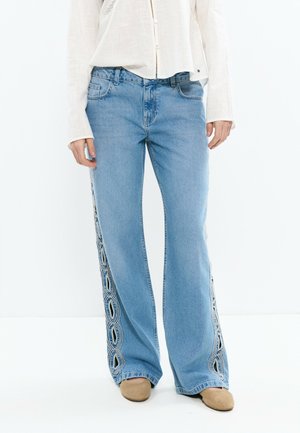 Femme portant un jean large bleu clair avec des découpes ovales brodées le long des coutures extérieures, un chemisier blanc et des chaussures beiges.