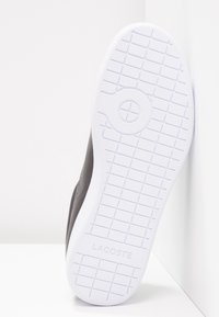 Svart sneaker med en slät läderövre, gummisula med ett rutmönster och präglad "LACOSTE"-logotyp på sulan.