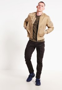 Chaqueta bomber beige con bolsillos con cremallera, superpuesta sobre una camisa de un verde oscuro y pantalones cargo negros, complementada con zapatillas deportivas azul marino.