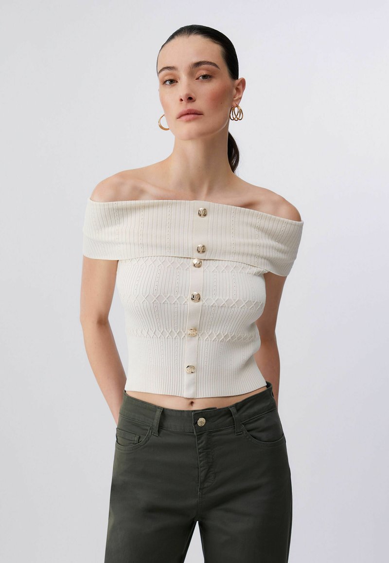 Femme aux cheveux foncés attachés en queue de cheval basse, portant un haut en maille blanc boutonné à épaules dénudées et un pantalon vert foncé taille haute, debout devant un fond uni.