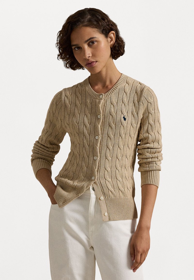 Cardigan en tricot beige avec un col rond, fermeture à boutons, poignets côtelés et un petit logo sur la poitrine gauche. Matériau léger et texturé.