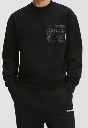Mann trägt schwarzen Pullover mit gemustertem schwarz-weißem Brusttaschenmuster und passende schwarze Hose mit weißem "Karl Lagerfeld"-Logo.