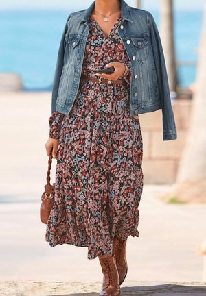 Femme portant une robe fleurie, une veste en jean posée sur les épaules, une ceinture marron, des bottes lacées, tenant un sac à poignée tressée marron et un smartphone.