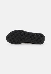Semelle de chaussure de sport noire avec une surface rugueuse et texturée présentant des motifs variés pour la traction et l'adhérence. Matière en caoutchouc durable.
