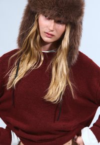Un pull bourgogne profond avec une texture douce, présentant une coupe ample et des manches longues. Assorti à un chapeau en fourrure marron, rehaussé de cordons de tirage noirs.
