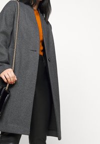 Šedý kabát s vzorem psí zub, střižený na míru, s jedním zapínacím knoflíkem, nošený přes oranžový žebrovaný svetr a černé kalhoty.