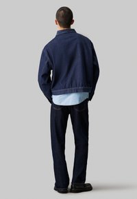 Giacca di jeans scura e jeans coordinati, con una camicia azzurra chiara sotto. L'outfit ha una vestibilità comoda con cuciture visibili.