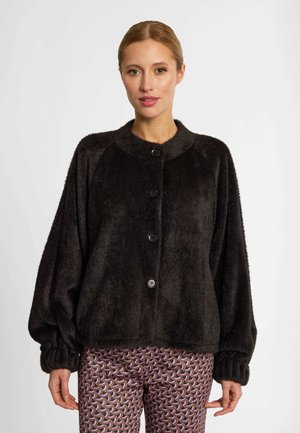 Vrouw draagt een zwart pluizig jasje met knopen en lange mouwen en een broek met patroon, staand tegen een witte achtergrond.
