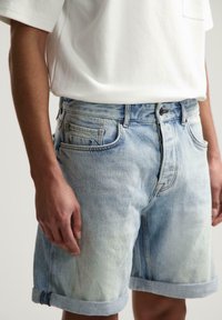 Hellblaue Denim-Shorts mit lockerer Passform, umgeschlagenen Säumen, fünf Taschen und sichtbarem Verblassen des Stoffes. Kombiniert mit einem weißen T-Shirt.