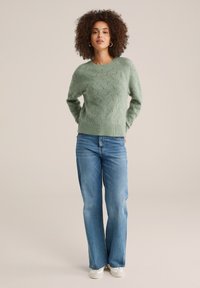 Pullover verde chiaro a maglia testurizzata con un motivo a onda, abbinato a jeans svasati blu e sneakers bianche. Il modello è in piedi contro uno sfondo neutro.