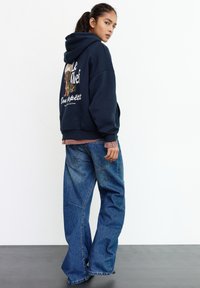 Felpa con cappuccio blu navy con grafica di un cane e testo sul retro, abbinata a jeans oversize in denim azzurro chiaro con grandi tasche e gambe ampie.