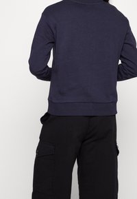 Sudadera de cuello redondo en azul marino, confeccionada en mezcla de algodón, con puños y dobladillo acanalados. Combinada con pantalones cargo negros que presentan bolsillos laterales con solapas.