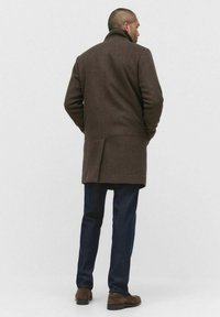 Bruine wollen jas met een hoge kraag, enkele split en achternaaddetail, gecombineerd met donkere jeans en bruine suède schoenen.