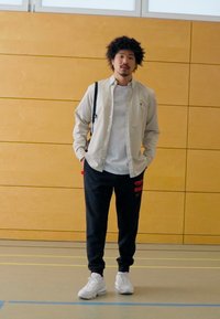 Ung mann iført lys beige skjorte over en hvit t-skjorte, marineblå joggebukser med rød tekst, og hvite sneakers, som står innendørs.