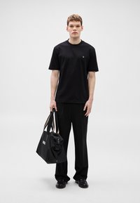 Μαύρο βαμβακερό t-shirt με στρογγυλή λαιμόκοψη, φορεμένο με μαύρο παντελόνι φαρδύ στο πόδι. Το μοντέλο κρατάει μια μαύρη τσάντα με την εκτύπωση "BOSS", που διαθέτει διπλές λαβές.