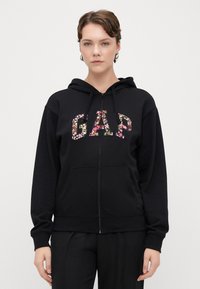 Črni zip-up dukserica z rožnatim logotipom "GAP", z kapuco na vrvico in sprednjimi žepi. Gladek material, sproščen kroj in rebrasti manšeti.