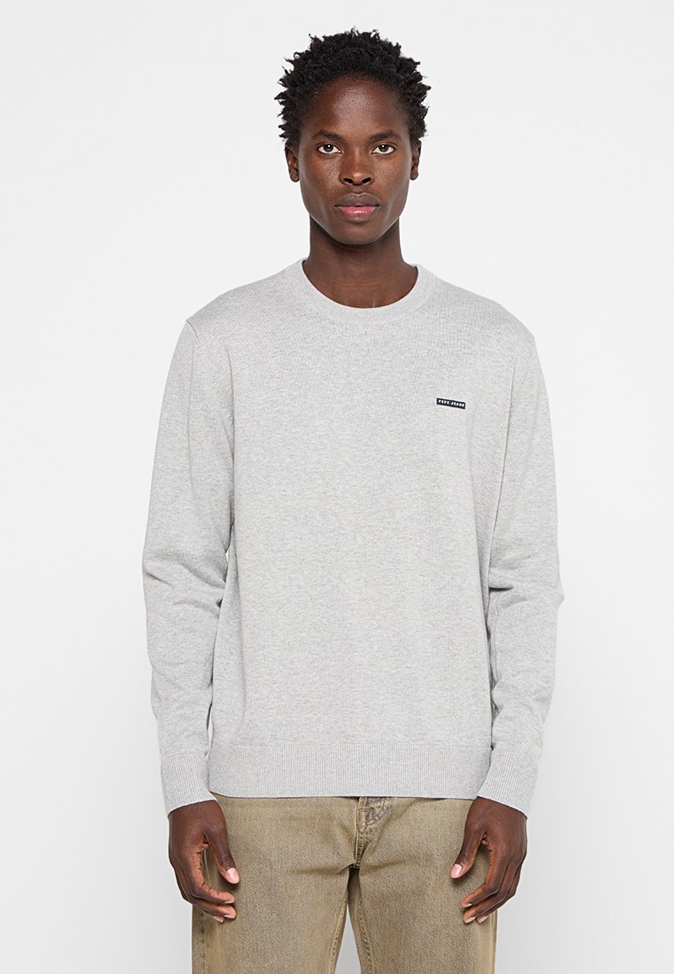 Pull gris à manches longues en tricot, avec un col rond et des poignets côtelés. Présente une petite écusson logo bleu foncé sur la poitrine.