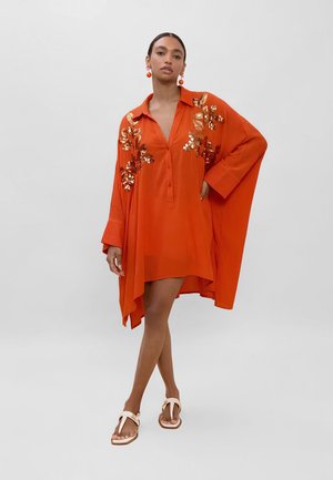 Donna in piedi che indossa un abito arancione ampio con dettagli floreali in paillettes sulle spalle, abbinato a orecchini arancioni e sandali crema.