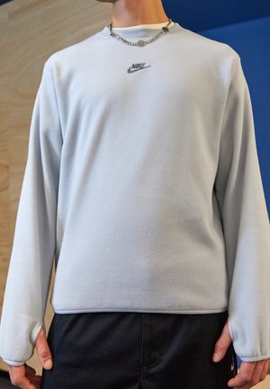 Person trägt einen hellgrauen Nike Langarm-Sweatshirt und schwarze Hose, steht vor einem hölzernen und blauen Hintergrund.