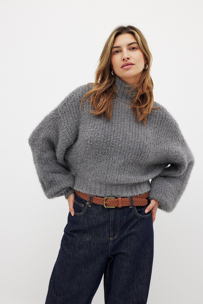 Grauer, grob gestrickter Pullover mit hohem Kragen und weiten Ärmeln, kombiniert mit einer dunklen Jeans und einem geflochtenen braunen Gürtel.
