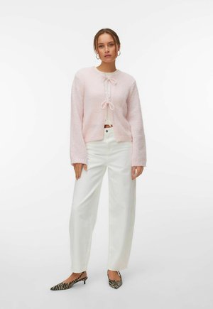 Femme portant un cardigan duveteux rose pâle noué avec des nœuds, un pantalon blanc à jambes larges et des chaussures à talons bas à motif zèbre, debout devant un fond blanc.