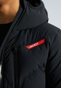 Veste matelassée noire avec capuche, arborant un logo rouge "JACKIT". Comprend une poche zippée, une texture matelassée et un design élégant.