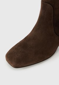 Brūni zamšādas potītes zābaki ar kvadrātu galu, gludu tekstūru un elegantu dizainu, kas izceļas ar sānu šuvju detaļām un nelielu papēdi.
