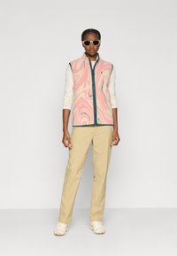 Γιλέκο πουλόβερ με μοτίβο ροζ και λευκής σπείρας, με φερμουάρ και τελειώματα σε τιρκουάζ. Φορεμένο πάνω από ένα μακρυμάνικο κρεμ τοπ και σε συνδυασμό με μπεζ παντελόνι.