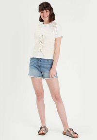 Cream crochet vest with a V-neck and button front, worn over a white t-shirt. Paired with distressed blue denim shorts and bronze sandals.