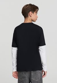 Czarny t-shirt z krótkim rękawem i okrągłym dekoltem, noszony na białej koszuli z długim rękawem, zestawiony z szarymi dżinsami. Koszula ma gładką fakturę.