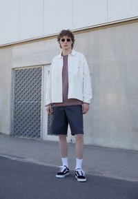 Lätt denimjacka i krämfärg, brun t-shirt, mörka shorts, vita strumpor och svarta sneakers med vita detaljer. Rund solglasögon bärs.