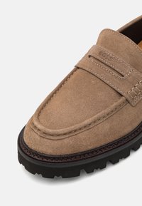 Mocassins en daim marron avec une surface texturée, présentant un détail cousu le long du bord et une semelle en caoutchouc noir robuste.