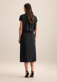 Schwarzes knielanges Kleid mit kurzen Ärmeln, elastischem Taillenbund und glattem Fabric. Tragen mit schwarzen High Heels. Ansicht von hinten.