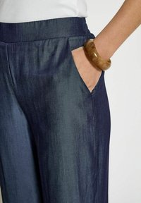 Pantalons larges bleu marine avec une texture lisse et un éclat subtil, dotés de poches latérales. Accompagnés d'un bracelet brillant marron au poignet.
