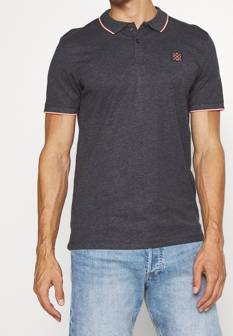 Homme portant un polo gris foncé avec des accents corail et un petit logo sur la poitrine, associé à un jean en denim bleu clair.